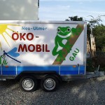 Ökomobil-Fahrzeuganhänger in der Seitenansicht. Darauf steht bunt "Neu-Ulmer Ökomobil" geschrieben sowei Frosch, Sonne und Libelle.