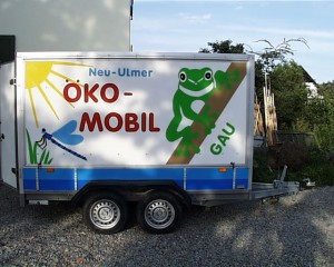 Ökomobil-Fahrzeuganhänger in der Seitenansicht. Darauf steht bunt "Neu-Ulmer Ökomobil" geschrieben sowei Frosch, Sonne und Libelle.